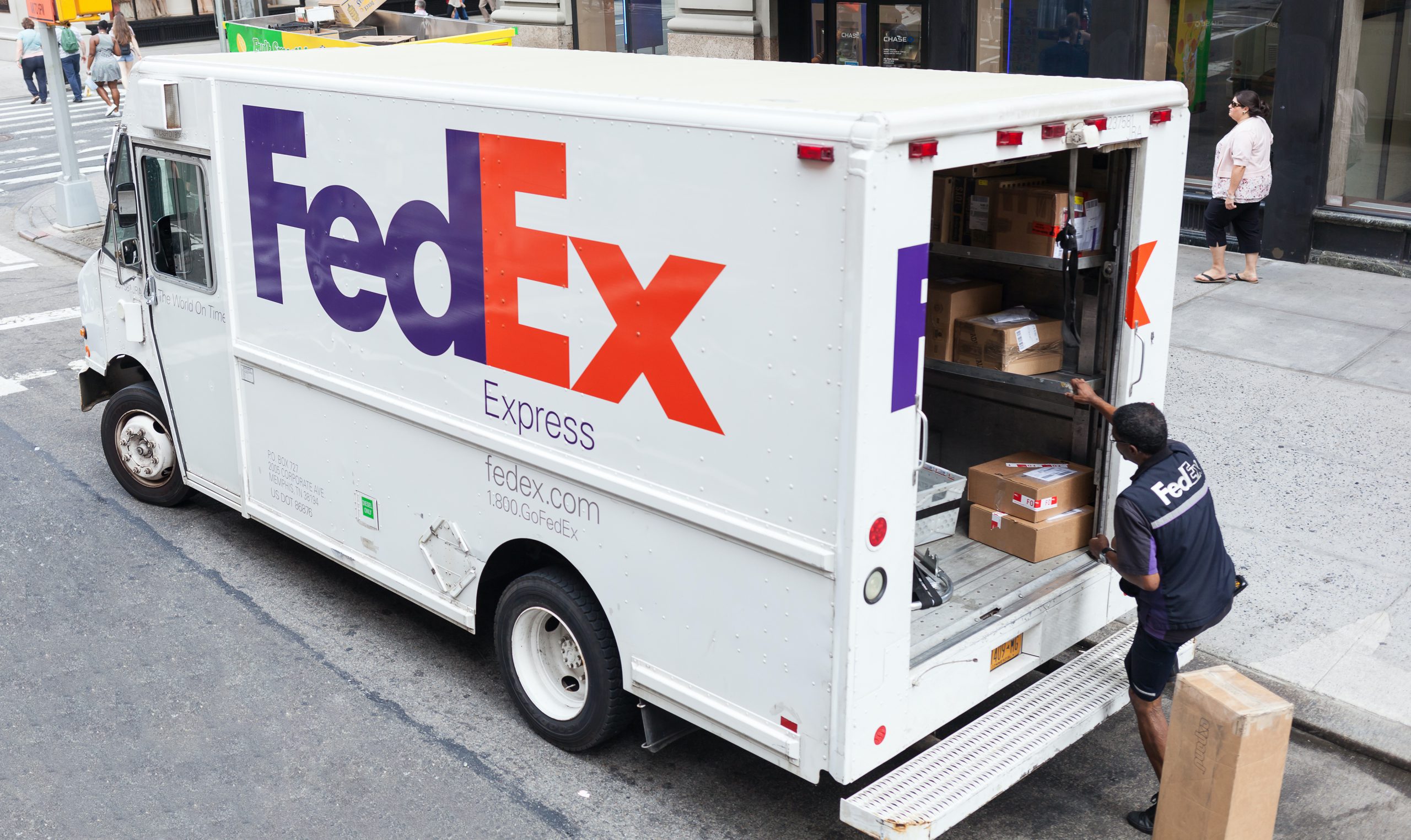 A FedEx worker reaches into the back of an open FedEx van to deliver a package outside a busy street corner